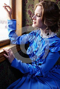 Young woman in blue vintage dress looking out the window in coup