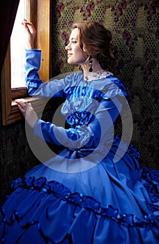 Young woman in blue vintage dress looking out the window in coup