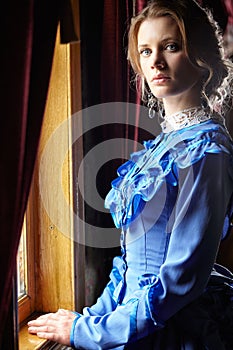 Young woman in blue vintage dress standing near window in coupe