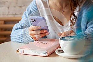 Young woman in blue warm sweater is reading a text message from mobile phone while drinking coffee