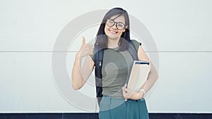 Young woman with books and backpack showing thumb up on grey background