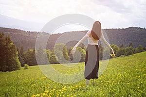 Young woman with a bouquet of flowers and wide open arms standing in the meadow. Forest and mountains in the background. Back view