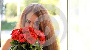 Young woman with a bouquet of red roses near an open window. A beautiful girl enjoys the aroma of flowers