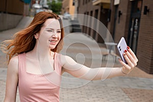 Young woman with braces on her teeth smiles and takes a selfie on a smartphone outdoors.