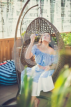 A young woman with bright makeup is sitting in a summer outdoor cafe in a hanging chair and blowing soap bubbles