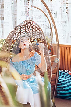 A young woman with bright makeup is sitting in a summer outdoor cafe in a hanging chair and blowing soap bubbles