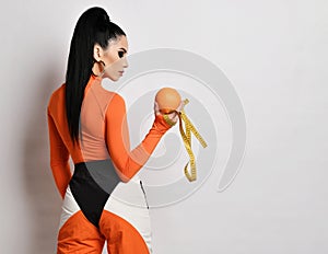 Young woman in bright orange sportswear and turtleneck stands back looking at fresh orange and measuring tape she holds