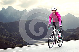 Young Woman in Bright Pink Jacket Riding Road Bicycle on Mountain Alpine Road. Healthy Lifestyle and Adventure Concept