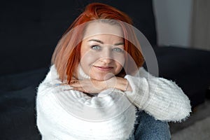 young woman with bright red hair sits on the floor at home in a white warm sweater