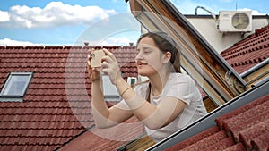 Young woman, with a bright smile, uses her smartphone to capture photos while looking out of an open attic window