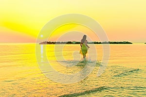 A young woman in a bright yellow dress meets the sunrise, joyfully runs through the sea water.