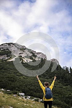Young woman in bright yellow sweater taking a rest during a hike standing with her arms raised high