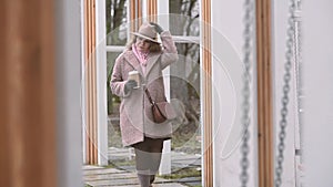 Young woman in a brown coat walking by swings with a cup of coffee