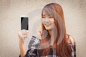 Young woman with brown hair smiling showing a blank smartphone screen standing on concrete wall background.
