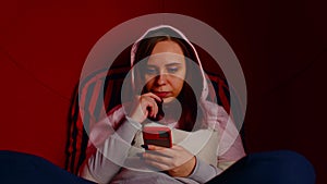Young woman browsing mobile phone and sitting in round chair. Female in hood using smartphone and resting in modern