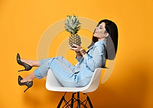 Young woman brunette in blue official suit and high-heeled shoes sitting in designer armchair with pineapple on yellow