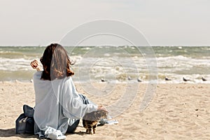 Young woman brunette in a light blue cardigan and jeans, with a backpack, sits on the beach and plays with a cat