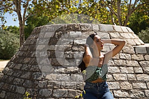 Young woman, brunette, slender, dressed in green t-shirt and jeans, leaning against a stone wall, hands in her hair relaxed.