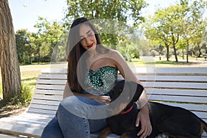 Young woman, brunette, slender, dressed in green t-shirt and jeans, sitting on a bench next to her dog, happy and in a loving