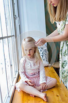 Young woman brushing pretty girl`s hair while sitting in front of window