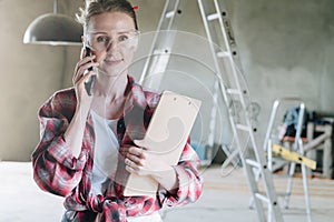 Young woman builder, architect, designer dressed in shirt and glasses is standing in workshop, talking on cell phone