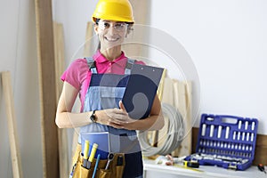 Young woman builder in protective overalls and hard hat smiling in workshop