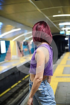 Young woman with burgundy pink maroon short hair, wearing purple top, light blue jeans and black sunglasses, waiting for train in