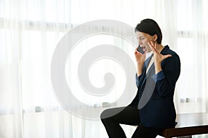 Young woman in a business casual suit sits at the edge of a table by the window of a hotel room and argues, gesturing with her