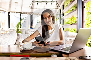 Young woman in a cafe reading a text message from her mobile phone. Female sitting at cafe table with laptop and using smart phone