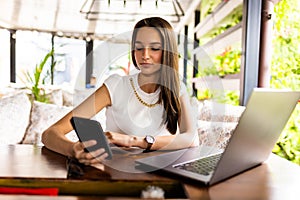 Young woman in a cafe reading a text message from her mobile phone. Female sitting at cafe table with laptop and using smart phone