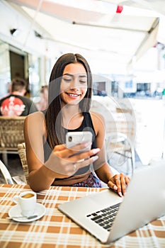 Young woman in a cafe reading a text message from her mobile phone. Latin female sitting at cafe table with laptop and using smart