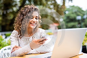 Young woman in a cafe reading a text message from her phone. Latin female sitting at cafe table with laptop and using smart phone.