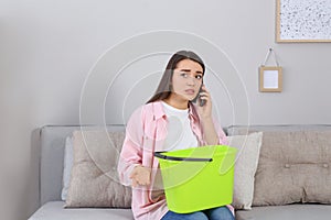 Young woman calling roof repair service while collecting leaking water from ceiling