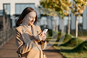 Young woman in camel color autumn coat walking on the street and using smartphone