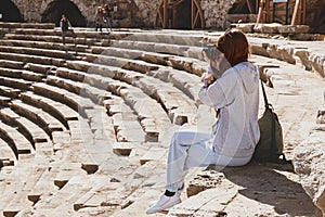 Young woman with camera. Caucasian tourist taking photo of ancient amphitheatre on sunny day. Soft blurred background