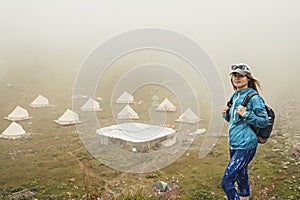 Young woman in cap and blue sportswear with backpack hiker standing in green mountain valley against the backdrop of tent camping