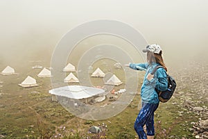 Young woman in cap and blue sportswear with backpack hiker standing in green mountain valley and pointing with hand at tent