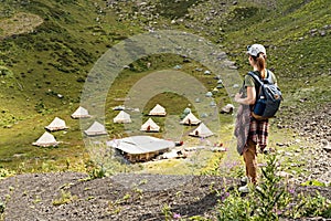 Young woman in cap and plaid shirt with backpack hiker standing in green mountain valley against the backdrop of tent camping in