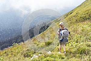 Young woman in cap and plaid shirt with big backpack hiking in green mountains against clouds in summer healthy active lifestyle,