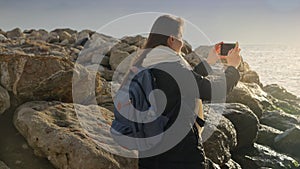 Young woman capturing the beauty of the winter sea by taking pictures with her smartphone while standing on the rocks.