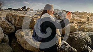 Young woman capturing the beauty of the winter sea by taking pictures with her smartphone while standing on the rocks. Ideal for