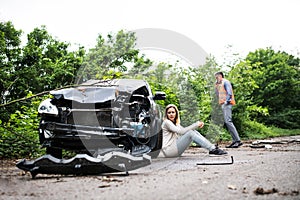 Young woman by the car after an accident and a man making a phone call.