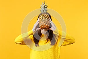 Young woman in casual clothes hiding, covering face with fresh ripe pineapple fruit isolated on yellow orange background