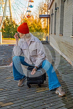 A young woman in casual clothes sits on a suitcase posing at the airport at the train station against the background of a brick