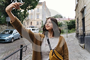 Young woman in casual fashionable outfit taking selfie while walking  in city street. Stylish hipster girl holding phone and