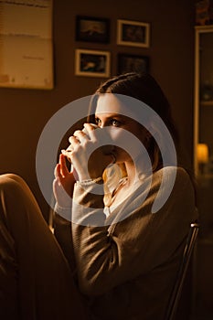 An young woman in casual home attire, sitting on a kitchen chair in the morning, drinking tea from a mug. Domestic comfort and