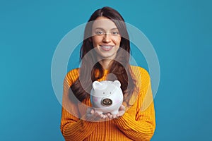 Young woman in casual orange sweater holding white piggy bank with lots of money