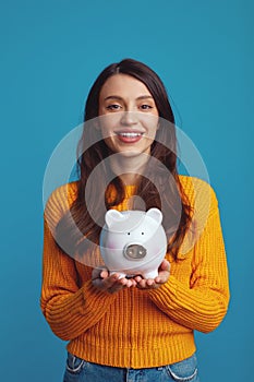 Young woman in casual orange sweater holding white piggy bank with lots of money