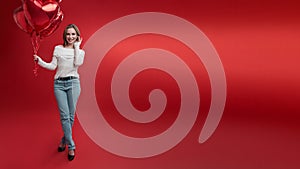 Young woman in casual style wear holds bunch of balloons on red background.