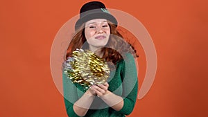 Young woman celebrating saint patrick`s day on orange wall in hat holding pom poms